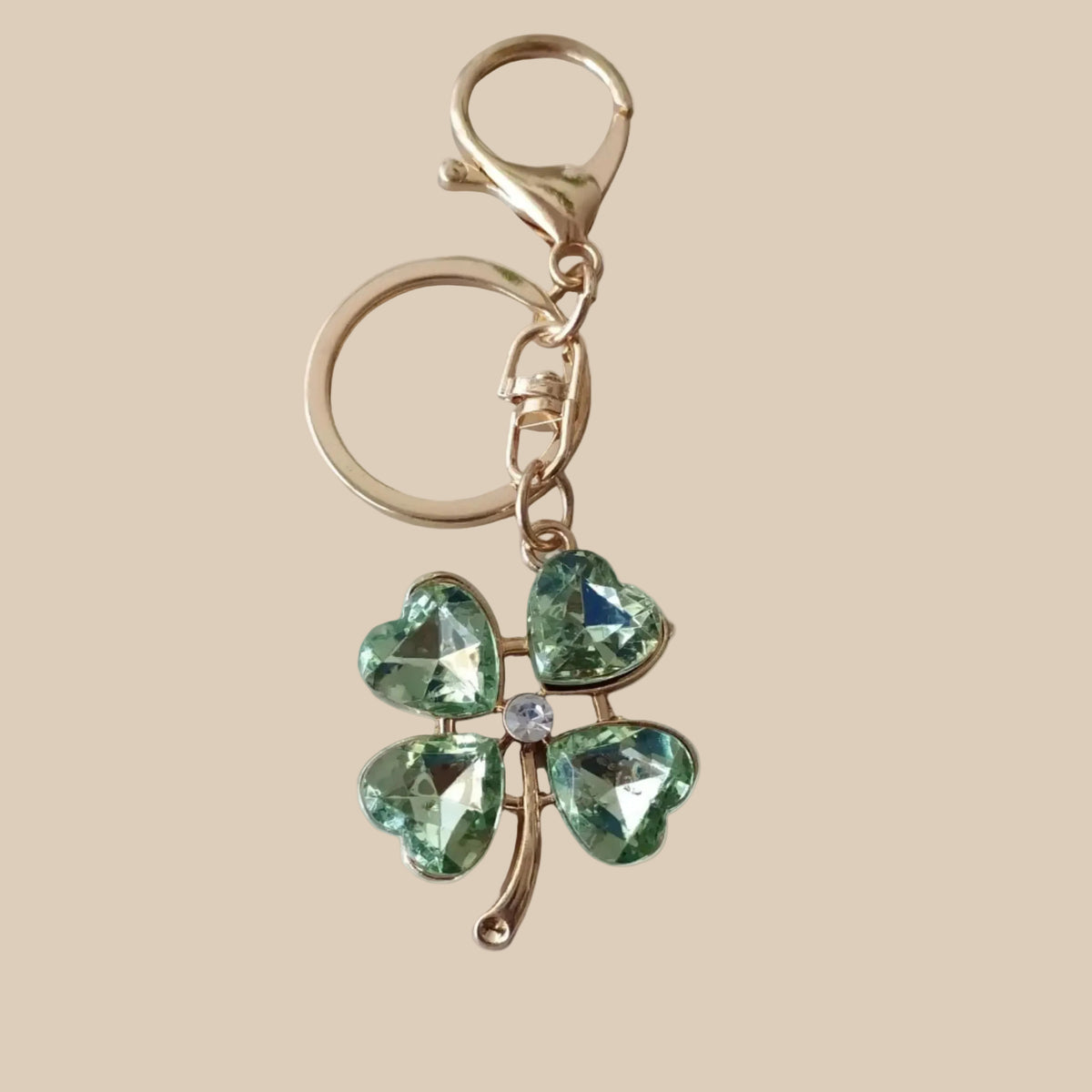 Bijou 3-en-1 Trèfle Émeraude One2K – vue produit fond sable, trèfle à quatre feuilles en cristal vert facetté, finitions dorées et cœur cristal blanc, anneau et mousqueton dorés