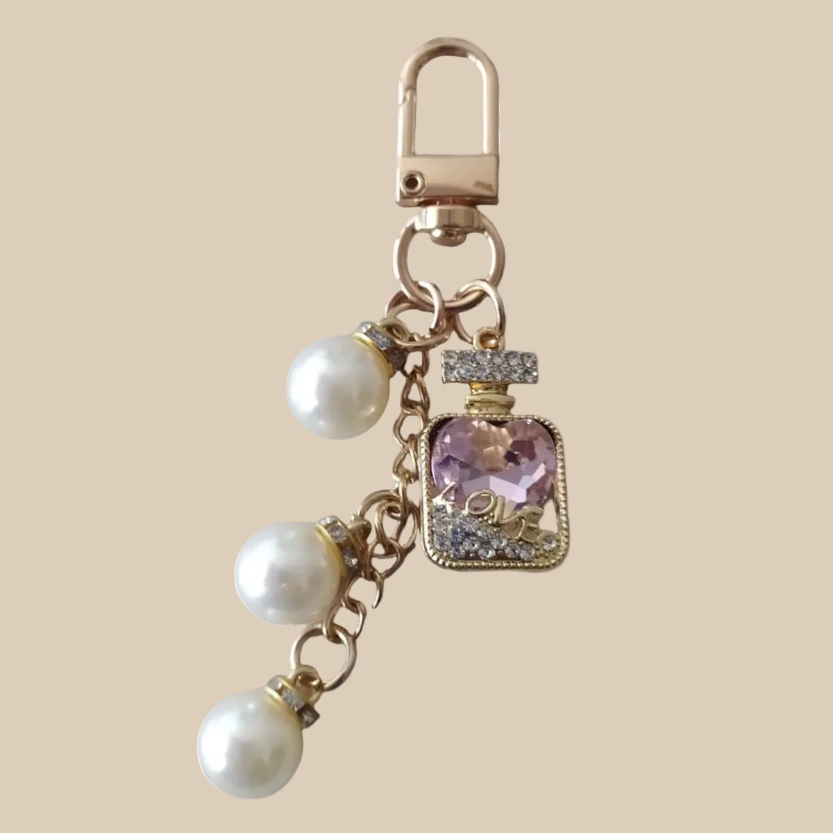 Bijou de sac téléphone 3-en-1 Élixir Rose One2K – vue présentation sur fond sable, flacon de parfum doré serti de strass avec cristal rose en forme de cœur gravé LOVE, perles nacrées blanches et mousqueton doré
