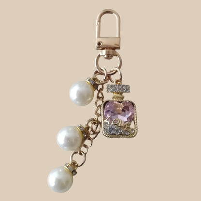 Bijou de sac téléphone 3-en-1 Élixir Rose One2K – vue présentation sur fond sable, flacon de parfum doré serti de strass avec cristal rose en forme de cœur gravé LOVE, perles nacrées blanches et mousqueton doré

