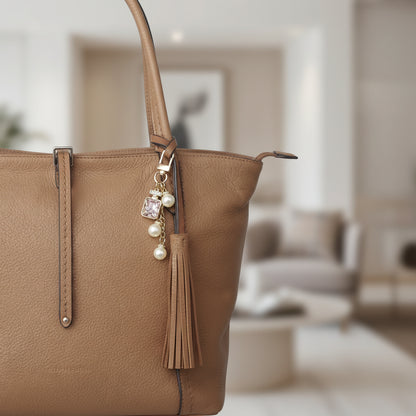 Bijou de sac téléphone 3-en-1 Élixir Rose One2K – situation d'usage accroché à un sac cabas en cuir camel avec pompon, flacon strass doré cristal rose et perles nacrées
