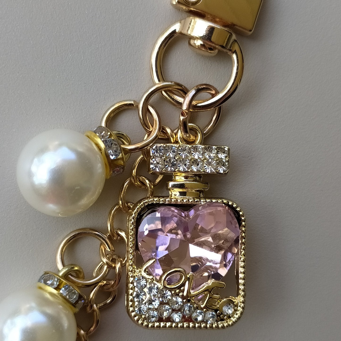 bijou de sac et de telephone 3 en 1  Elixir Rose One2K - gros plan sur fond beige - flacon de parfum doré migniature avec détails en strass et cristal rose en forme de coeur gravé LOVE, chainette ornée de perles nacrées