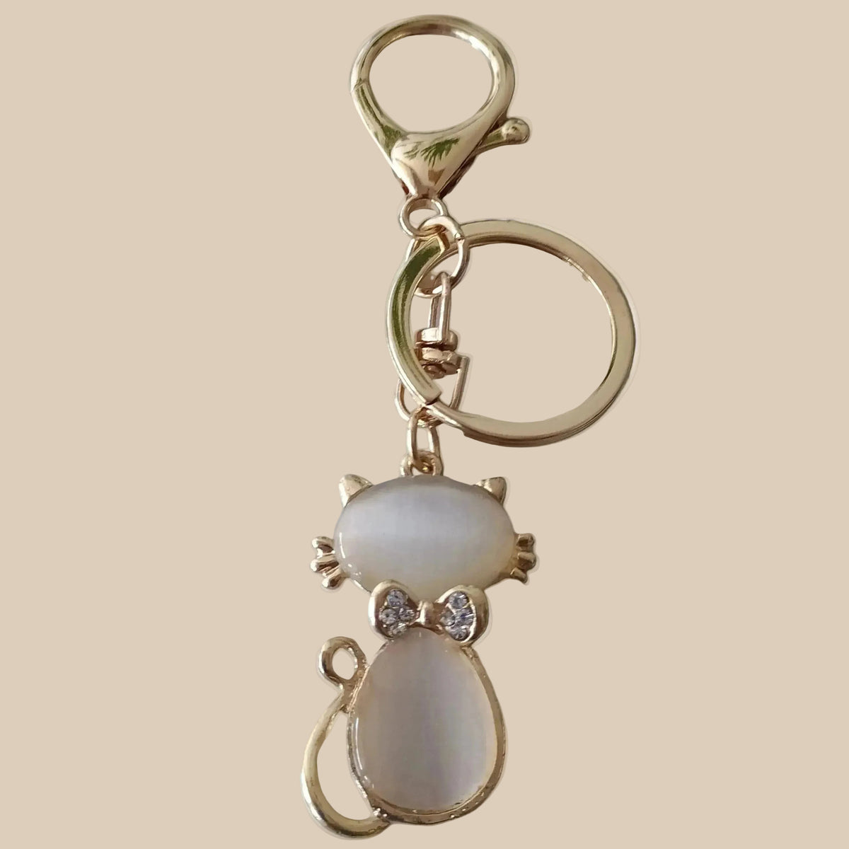 Bijou 3-en-1 Félin Poudré One2K – vue produit fond sable, chat en émail nacré blanc et contours dorés avec nœud strass, anneau et mousqueton dorés