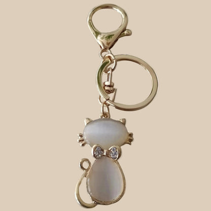 Bijou 3-en-1 Félin Poudré One2K – vue produit fond sable, chat en émail nacré blanc et contours dorés avec nœud strass, anneau et mousqueton dorés