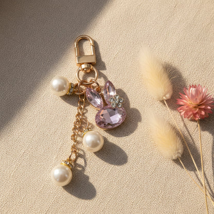 Bijou de sac téléphone 3-en-1 Lapin de Cristal One2K – photo ambiance sur tissu beige avec fleurs séchées crème et rose, lapin cristal rose, perles nacrées et chaîne dorée
