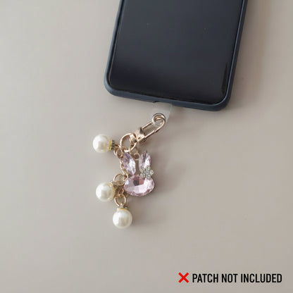 Bijou de téléphone 3 en 1 Lapin de Cristal One2K - silouhette de lapin en cristal rose au contour doré avec petite fleur cristal blanc, perles nacrés tombantes sur une chaine  dorée - situation clipé au téléphone sur un fond beige