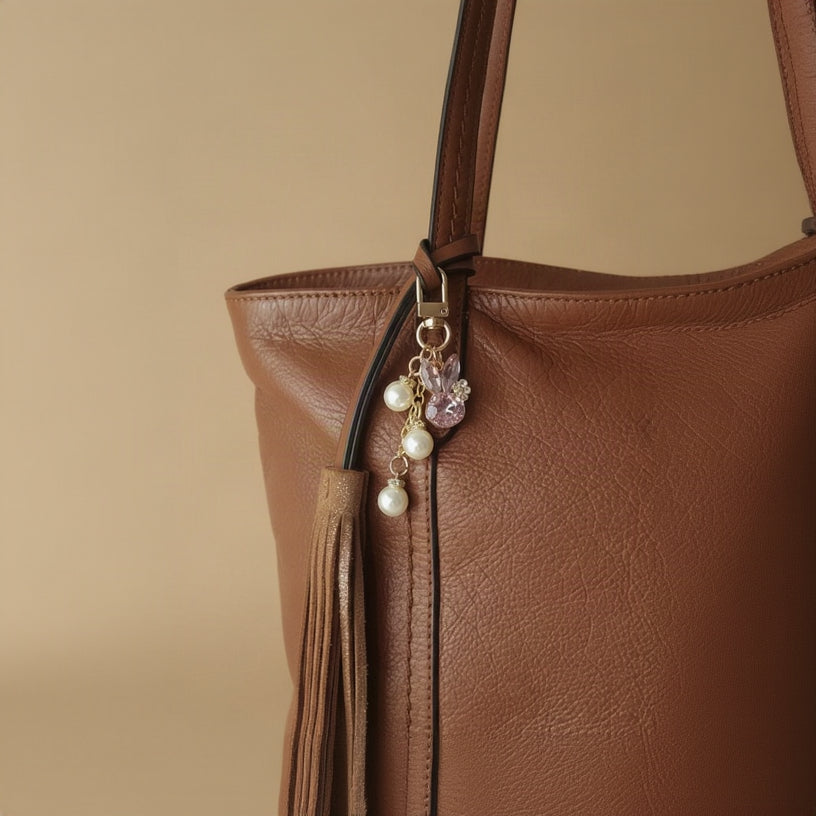 Bijou de sac téléphone 3-en-1 Lapin de Cristal One2K – situation d'usage accroché à un grand sac cabas en cuir camel avec pompon, lapin cristal rose et perles nacrées
