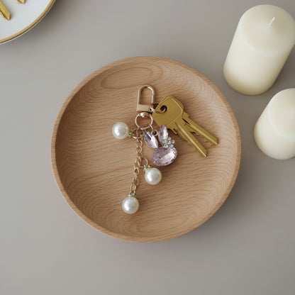 Bijou porte-clés Lapin de Cristal One2K utilisé comme porte-clés – lapin formé de cristaux rose et marquise transparents avec strass, chainette dorée ornée de perles nacrées blanches, accroché à un trousseau de clés posé sur un plateau en bois, bougies blanches en arrière-plan