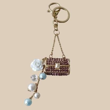 Bijou de sac téléphone 3-en-1 Mini Sac Pourpre One2K – vue présentation sur fond sable, mini sac orné de strass pourpres, chaîne dorée, rose blanche et perles