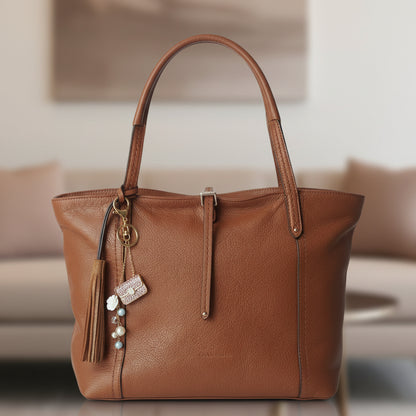 Bijou de sac téléphone 3-en-1 Mini Sac Pourpre One2K – situation d'usage accroché à un sac en cuir camel avec pompon, décoration florale dorée en arrière-plan