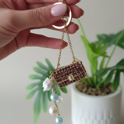 Bijou de sac téléphone 3-en-1 Mini Sac Pourpre One2K – photo dimensions tenu en main devant une plante verte, mini sac strass pourpre avec perles et fleur blanche