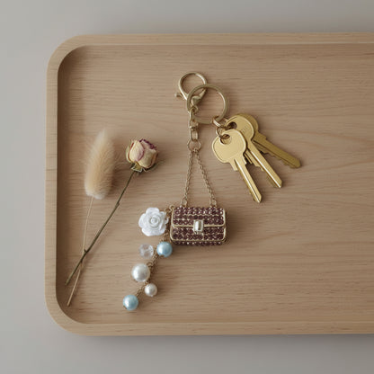Bijou porte-clés Mini Sac Pourpre One2K utilisé comme porte-clés – mini sac recouvert de strass pourpres, chaîne dorée, rose blanche en nacre et perles bleues et nacrées, accroché à un trousseau de clés posé sur un plateau en bois avec une rose séchée