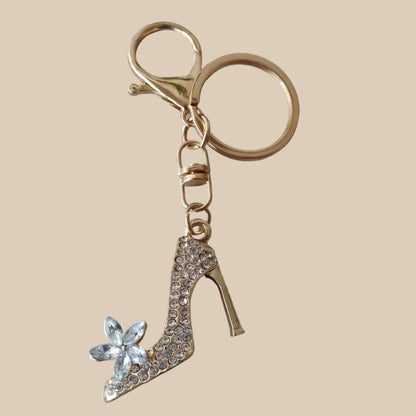 Bijou de sac téléphone 3-en-1 Soulier de Cristal One2K – vue présentation sur fond sable, escarpin doré serti de strass transparents avec fleur en cristal, anneau et mousqueton dorés
