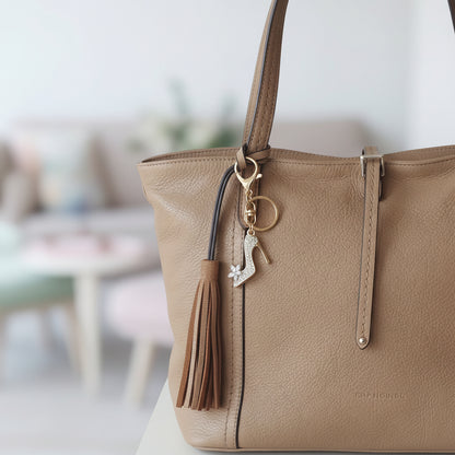 Bijou de sac téléphone 3-en-1 Soulier de Cristal One2K – situation d'usage accroché à un sac cabas en cuir camel avec pompon, décoration florale séchée en arrière-plan
