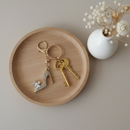 Bijou porte-clés Soulier de Cristal One2K utilisé comme porte-clés – escarpin doré serti de strass transparents avec fleur en cristal marquise, accroché à un trousseau de clés posé sur un plateau en bois, petit vase blanc avec fleurs séchées en arrière-plan
