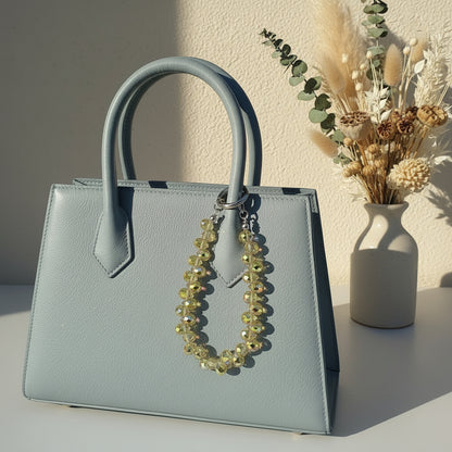 Bijou 2-en-1 Dragonne de Poignet & Bijou de Sac Éclat Boréal reflets solaires One2K – situation d'usage accroché à un sac bleu gris structuré, perles de cristal facettées aux reflets dorés et irisés solaires, fermoir anneau argenté