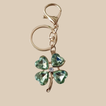 Bijou 3-en-1 Trèfle Émeraude One2K – vue produit fond sable, trèfle à quatre feuilles en cristal vert facetté, finitions dorées et cœur cristal blanc, anneau et mousqueton dorés