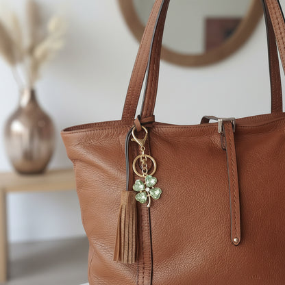 Bijou de sac et de telephone Trèfle Émeraude One2K clipé en bijou de sac sur un sac à main caramel en cuir, fleurs séchées en arrière-plan
