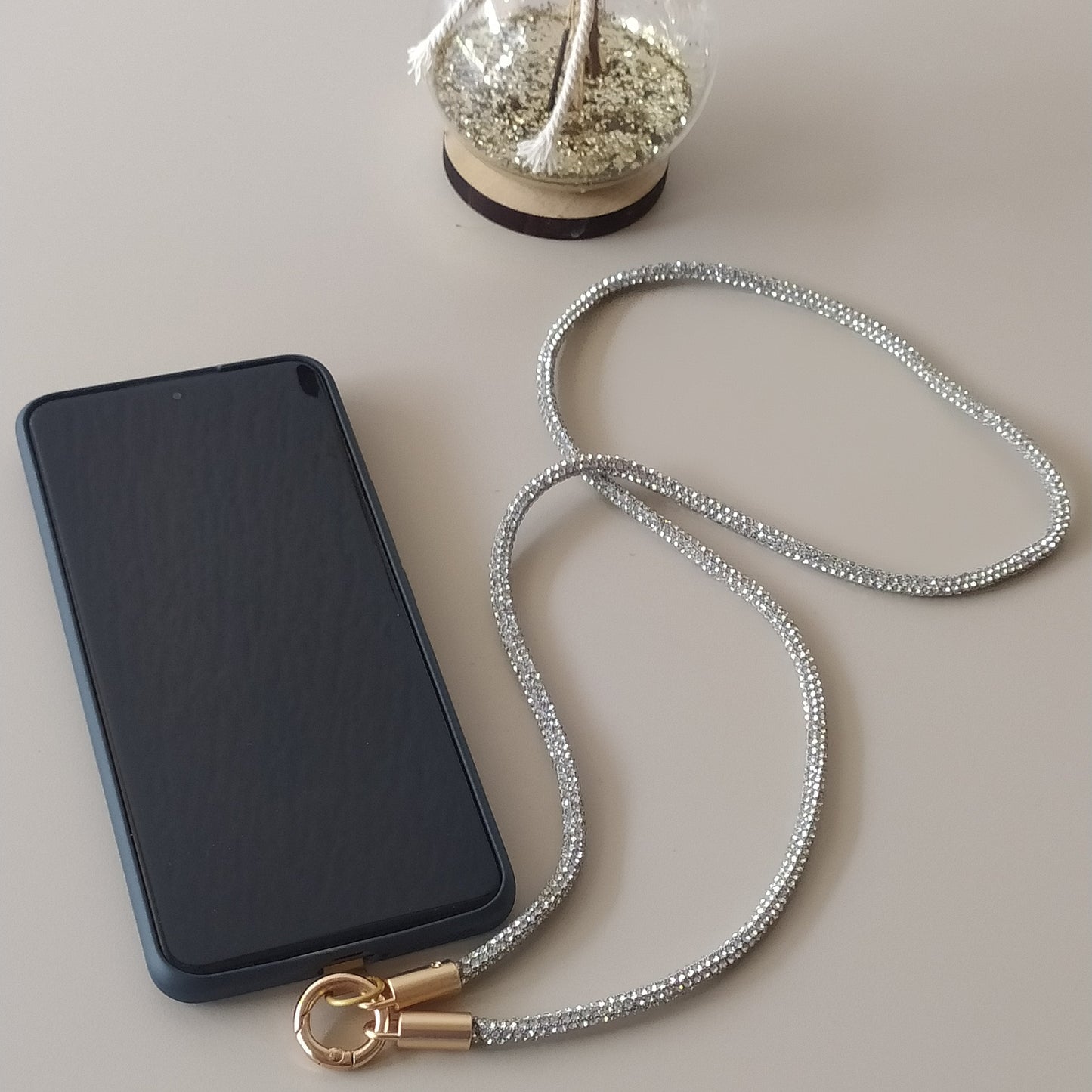 Collier téléphone infini strass One2K version argent – situation d'usage accroché à un smartphone noir sur fond gris, corde strass argentée à reflets scintillants et mousqueton doré
