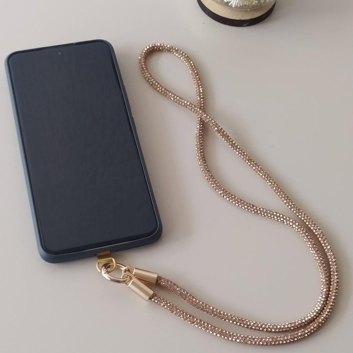 Collier téléphone infini strass One2K version or – situation d'usage accroché à un smartphone noir sur fond gris, corde strass dorée champagne à reflets scintillants et mousqueton doré

