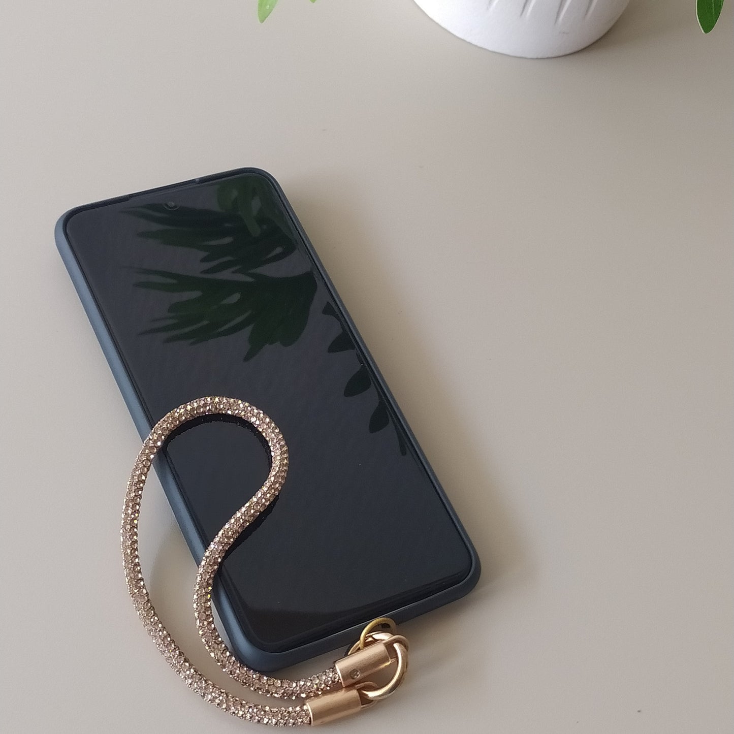 Dragonne poignet téléphone infini strass  One2K version or – situation d'usage accroché à un smartphone noir sur fond beige, corde tubulaire strass dorée champagne scintillante et fermoir doré

